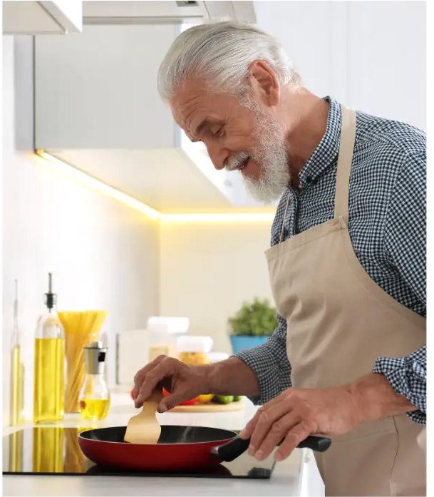 Older man in an apron stirring food in a red frying pan on the stove